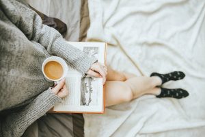 Girl reading a book with a cup of coffee. She has a green top, and pale skin. This photo is shot from the sky looking down, you cann see her torso and legs. She's sitting on her bed. 