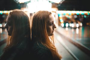 Woman standing outside, lights behind her blurry. Leaning against window reflecting her red hair.