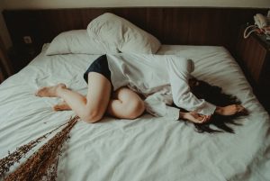 woman with brown hair laying across a white bed, wearing white long sleeve top and blue shorts, looking exhausted.