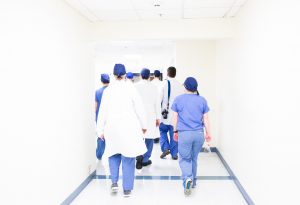 Doctors in white coats and pale blue scrubs with royal blue scrub hats walk away down the hall