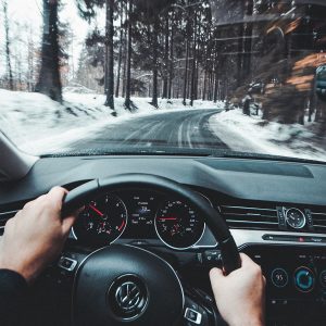 mans hands on the black steering wheel of a black car driving over a cleared road in snowy conditions.