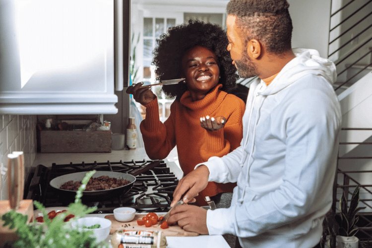 Woman at stove holding spoon for man to taste cooking