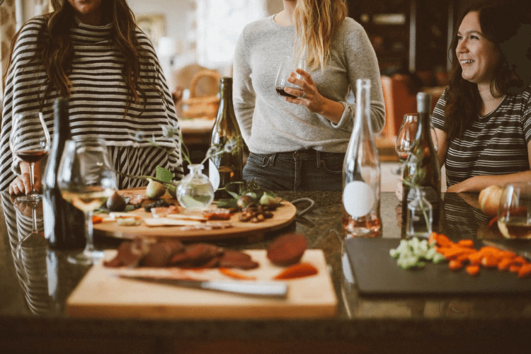 3 women standing around kitchen island that has food and wine on it